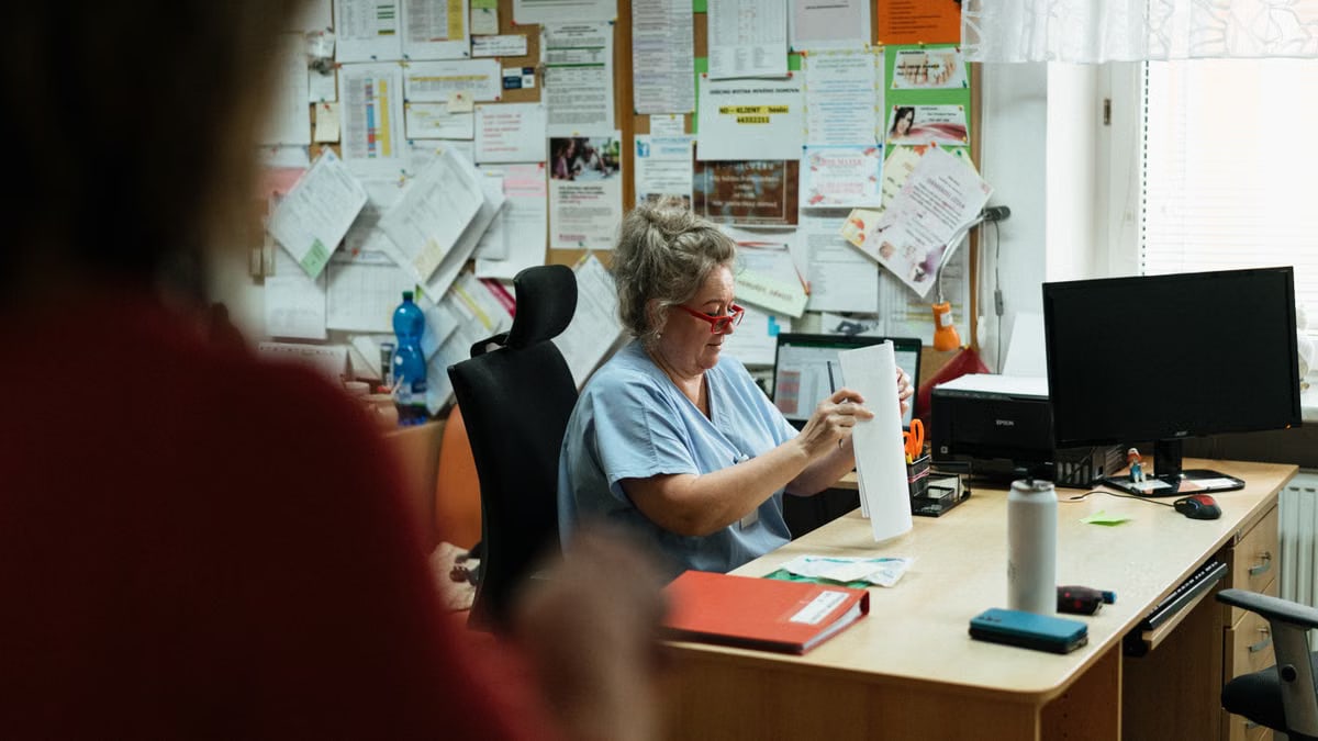 A senior caregiver in blue scrubs sorting paperwork at a hospital office desk.