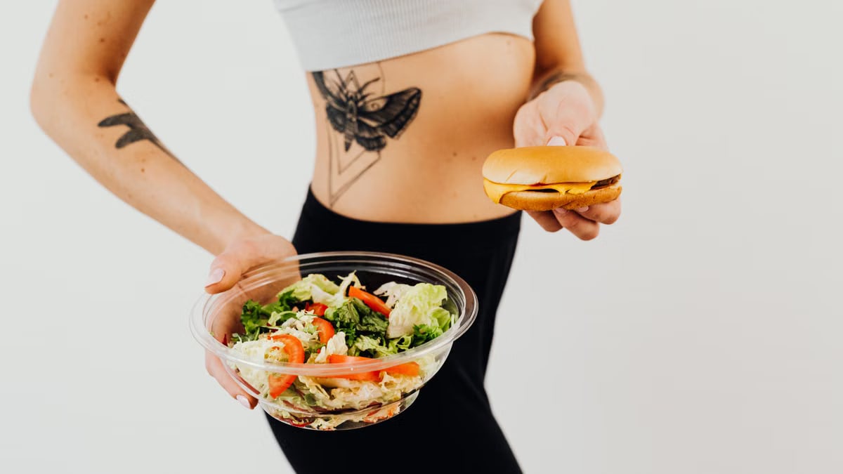 A woman with a tattoo holding a healthy vegetable salad and a cheeseburger, representing food choices.
